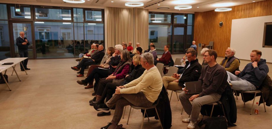 Menschen sitzen in einem Sitzungssaal in Reihen angeordnet. Vorne steht eine Person mit Mikrofon.