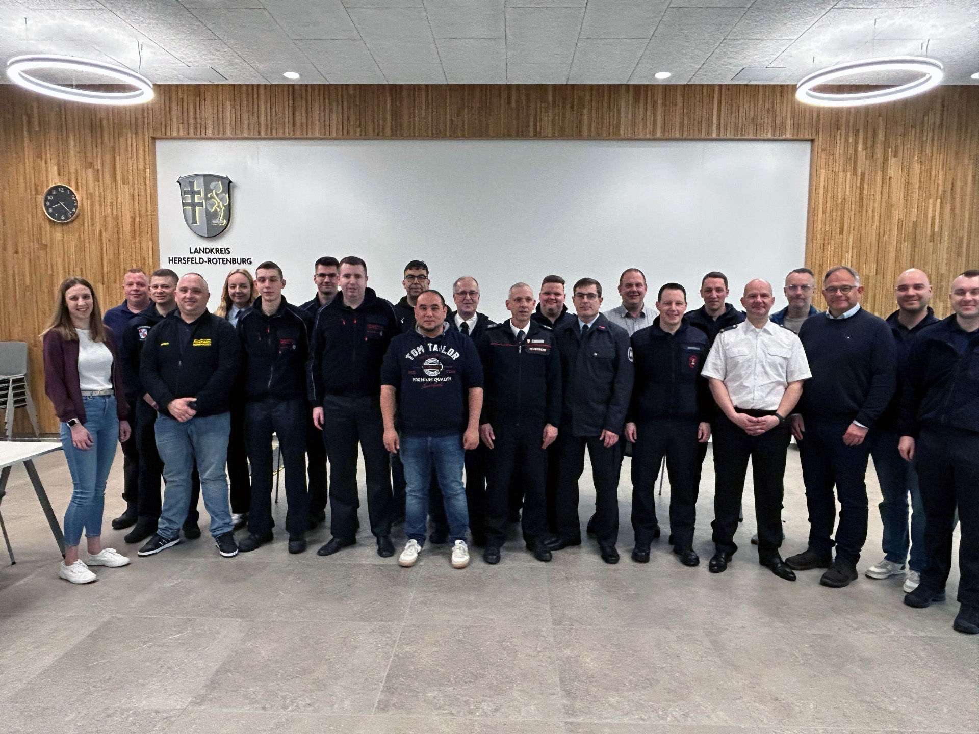 Eine Gruppe von etwa zwanzig Personen steht nebeneinander in einem modernen Sitzungssaal vor einer Wand mit dem Wappen und der Aufschrift „Landkreis Hersfeld-Rotenburg“. Einige tragen Feuerwehr-Uniformen, andere Freizeitkleidung. Links stehen zwei Frauen, die übrigen Personen sind überwiegend Männer. Über der Gruppe hängen runde Deckenleuchten, links an der Wand ist eine Uhr zu sehen.