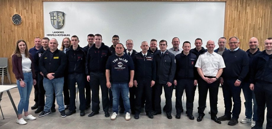Eine Gruppe von etwa zwanzig Personen steht nebeneinander in einem modernen Sitzungssaal vor einer Wand mit dem Wappen und der Aufschrift „Landkreis Hersfeld-Rotenburg“. Einige tragen Feuerwehr-Uniformen, andere Freizeitkleidung. Links stehen zwei Frauen, die übrigen Personen sind überwiegend Männer. Über der Gruppe hängen runde Deckenleuchten, links an der Wand ist eine Uhr zu sehen.