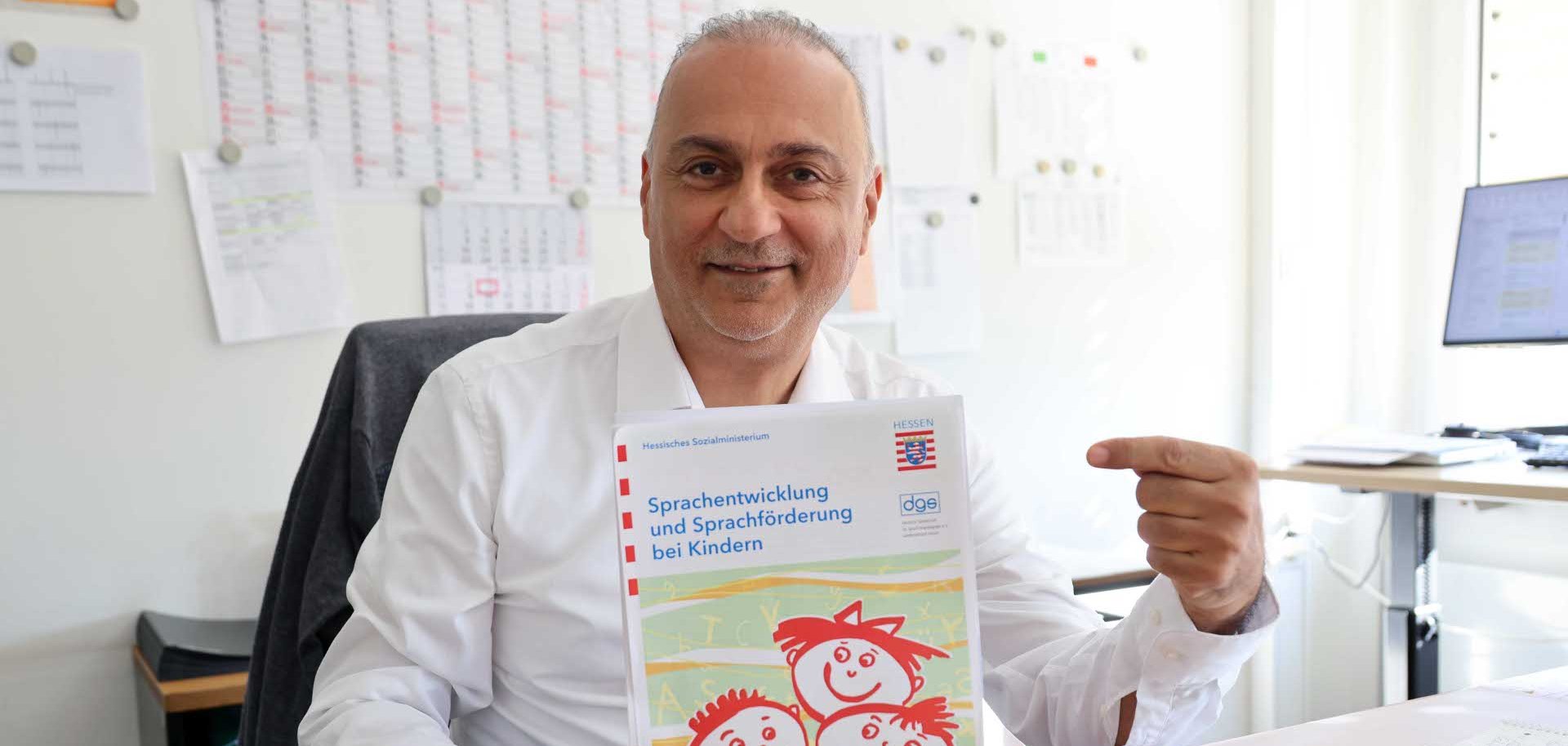 Auf dem Foto ist ein Mann in einem weißen Hemd zu sehen, der eine Zeitschrift in den Händen hält. Im Hintergrund ist zudem ein Kalender sowie ein Computerbildschirm zu sehen.