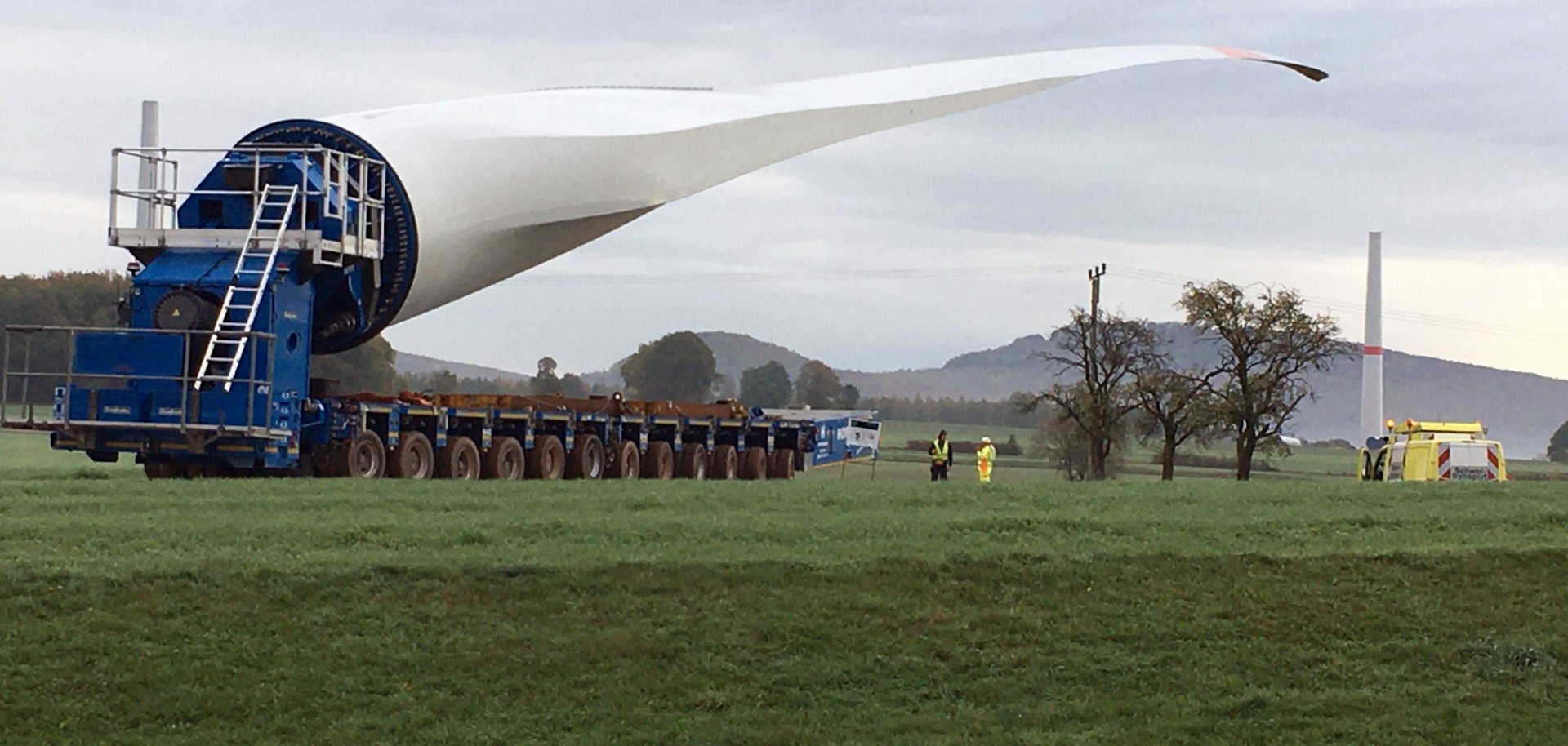 Ein sehr langes Rotorblatt einer Windkraftanlage wird auf einem speziellen Schwerlasttransporter über eine Wiese transportiert. Zwei Arbeiter stehen daneben, während im Hintergrund ein Turm einer Windkraftanlage und einige Bäume unter einem bewölkten Himmel zu sehen sind.