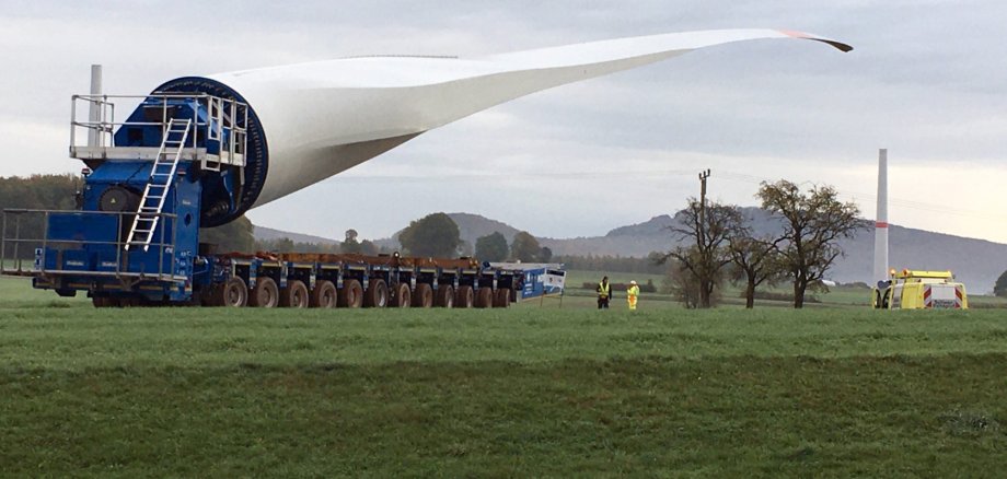 Ein sehr langes Rotorblatt einer Windkraftanlage wird auf einem speziellen Schwerlasttransporter über eine Wiese transportiert. Zwei Arbeiter stehen daneben, während im Hintergrund ein Turm einer Windkraftanlage und einige Bäume unter einem bewölkten Himmel zu sehen sind.