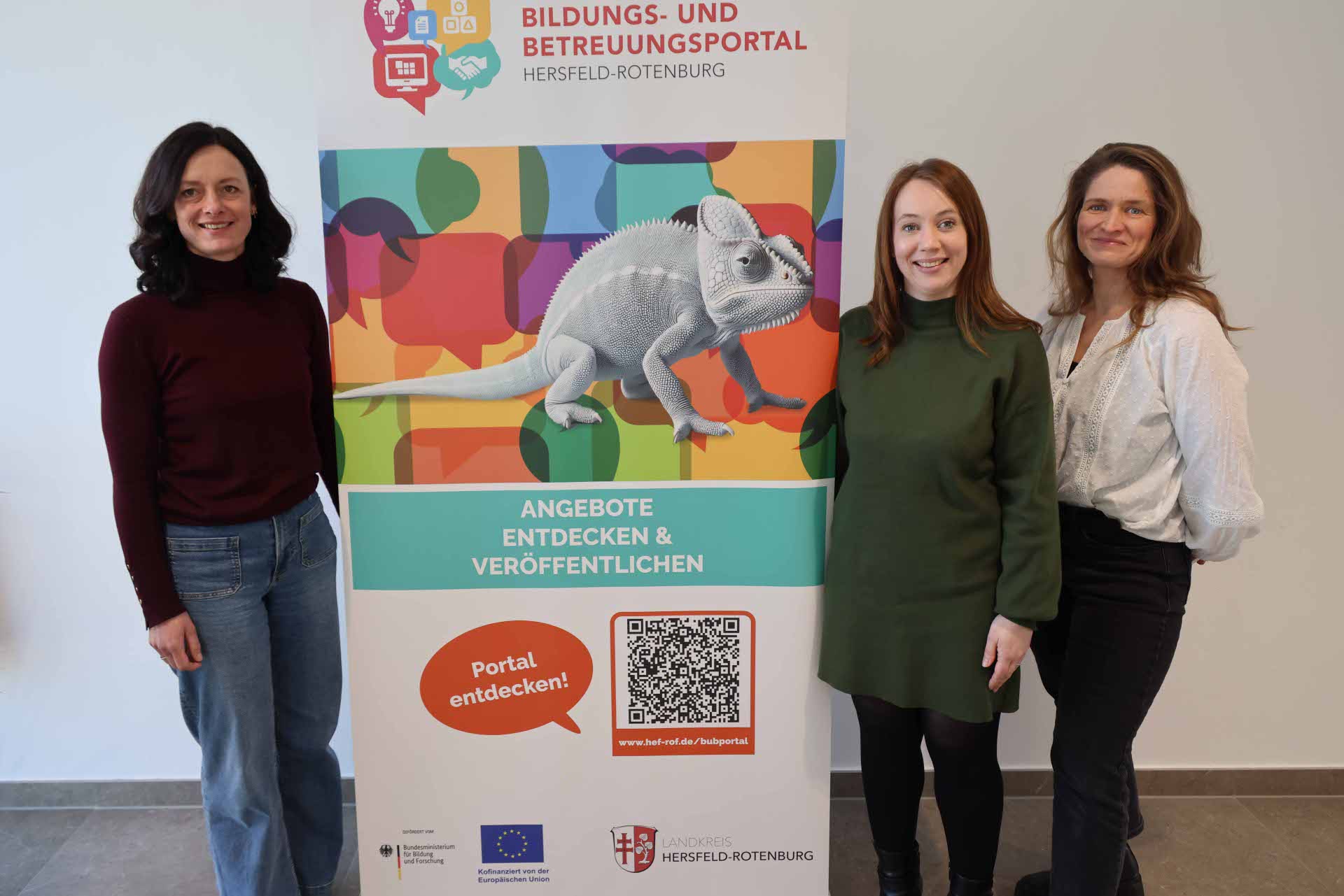 Drei Frauen stehen nebeneinander vor einem bunten Roll-up zum Bildungs- und Betreuungsportal des Landkreises Hersfeld-Rotenburg und lächeln in die Kamera.