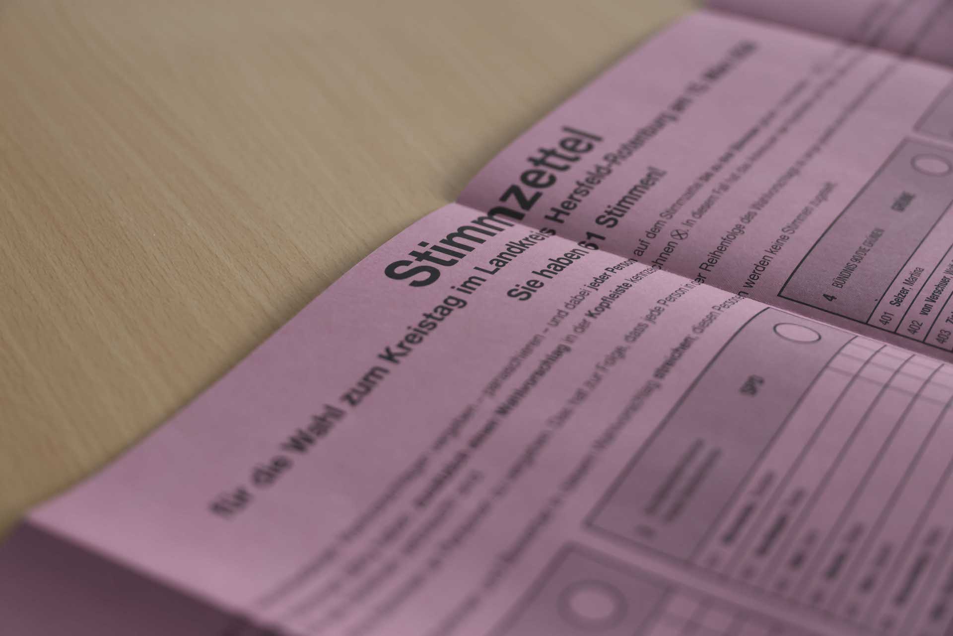 Nahaufnahme eines rosafarbenen Wahlzettels mit der Überschrift „Stimmzettel“ für die Wahl zum Kreistag im Landkreis Hersfeld-Rotenburg, der auf einem hellen Holztisch liegt; Teile der Kandidatenliste und Auswahlkreise sind sichtbar, der Hintergrund ist unscharf.