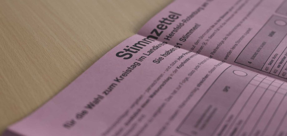 Nahaufnahme eines rosafarbenen Wahlzettels mit der Überschrift „Stimmzettel“ für die Wahl zum Kreistag im Landkreis Hersfeld-Rotenburg, der auf einem hellen Holztisch liegt; Teile der Kandidatenliste und Auswahlkreise sind sichtbar, der Hintergrund ist unscharf.