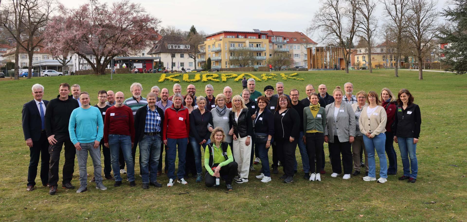 Eine Gruppe von Menschen steht im Bad Hersfelder Kurpark. Im Hintergrund sieht man gelbe Blumen, die das Wort "Kurpark" bilden.