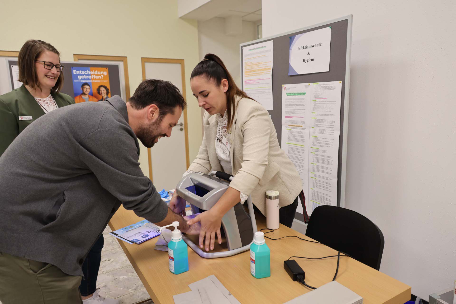 Eine Frau steht an der Station Infektionsschutz & Hygiene. Ein Mann legt seine Hände in ein Gerät. 