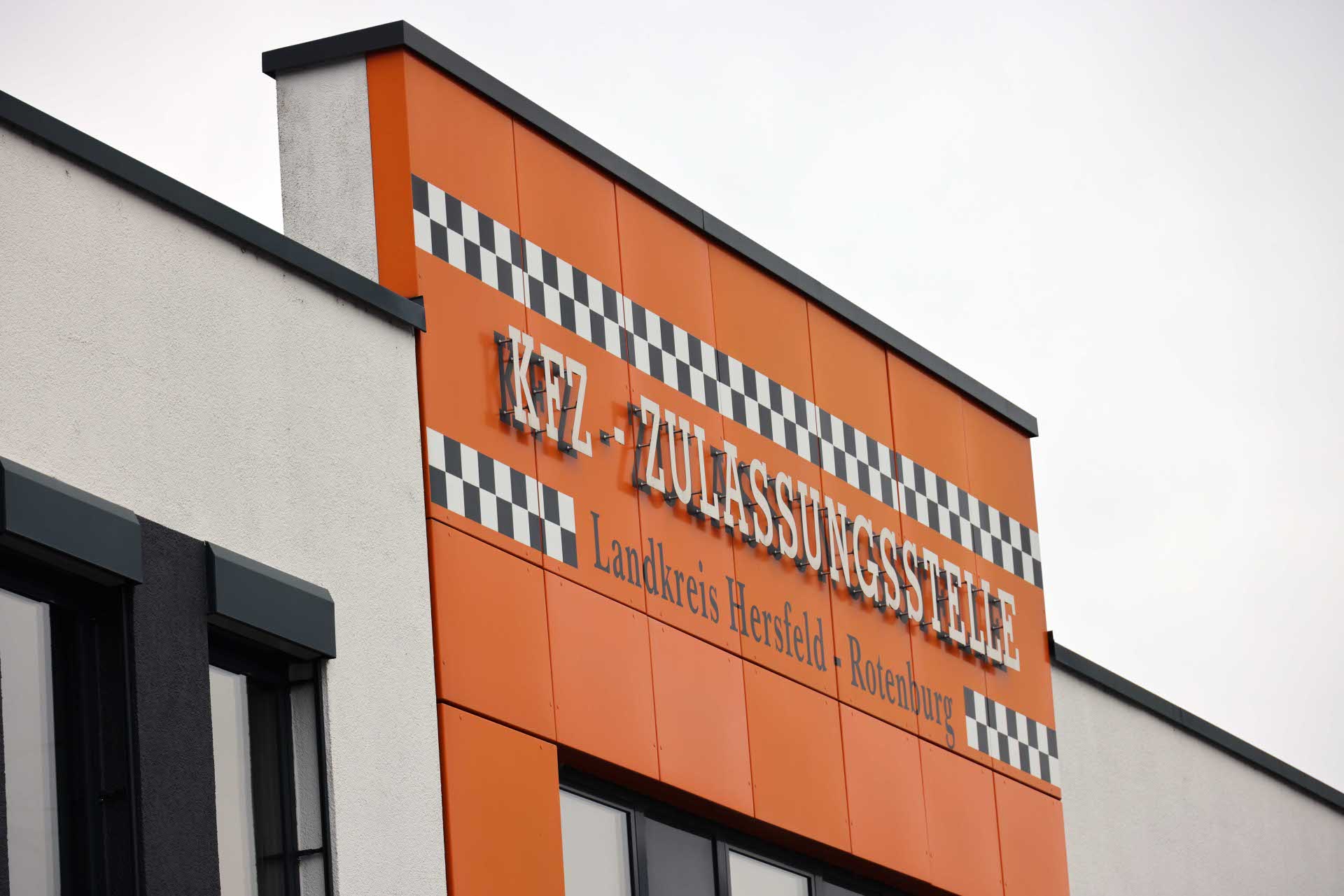 Gebäudefassade mit orangefarbener Verkleidung und dem Schriftzug ‚KFZ-Zulassungsstelle – Landkreis Hersfeld-Rotenburg‘ sowie schwarz-weißem Karomuster über den Fenstern.
