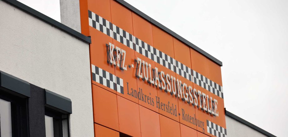 Gebäudefassade mit orangefarbener Verkleidung und dem Schriftzug ‚KFZ-Zulassungsstelle – Landkreis Hersfeld-Rotenburg‘ sowie schwarz-weißem Karomuster über den Fenstern.