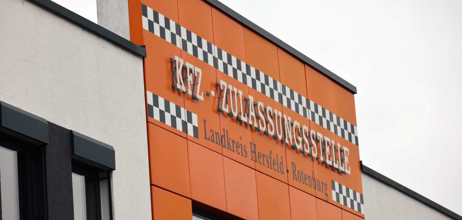 Gebäudefassade mit orangefarbener Verkleidung und dem Schriftzug ‚KFZ-Zulassungsstelle – Landkreis Hersfeld-Rotenburg‘ sowie schwarz-weißem Karomuster über den Fenstern.