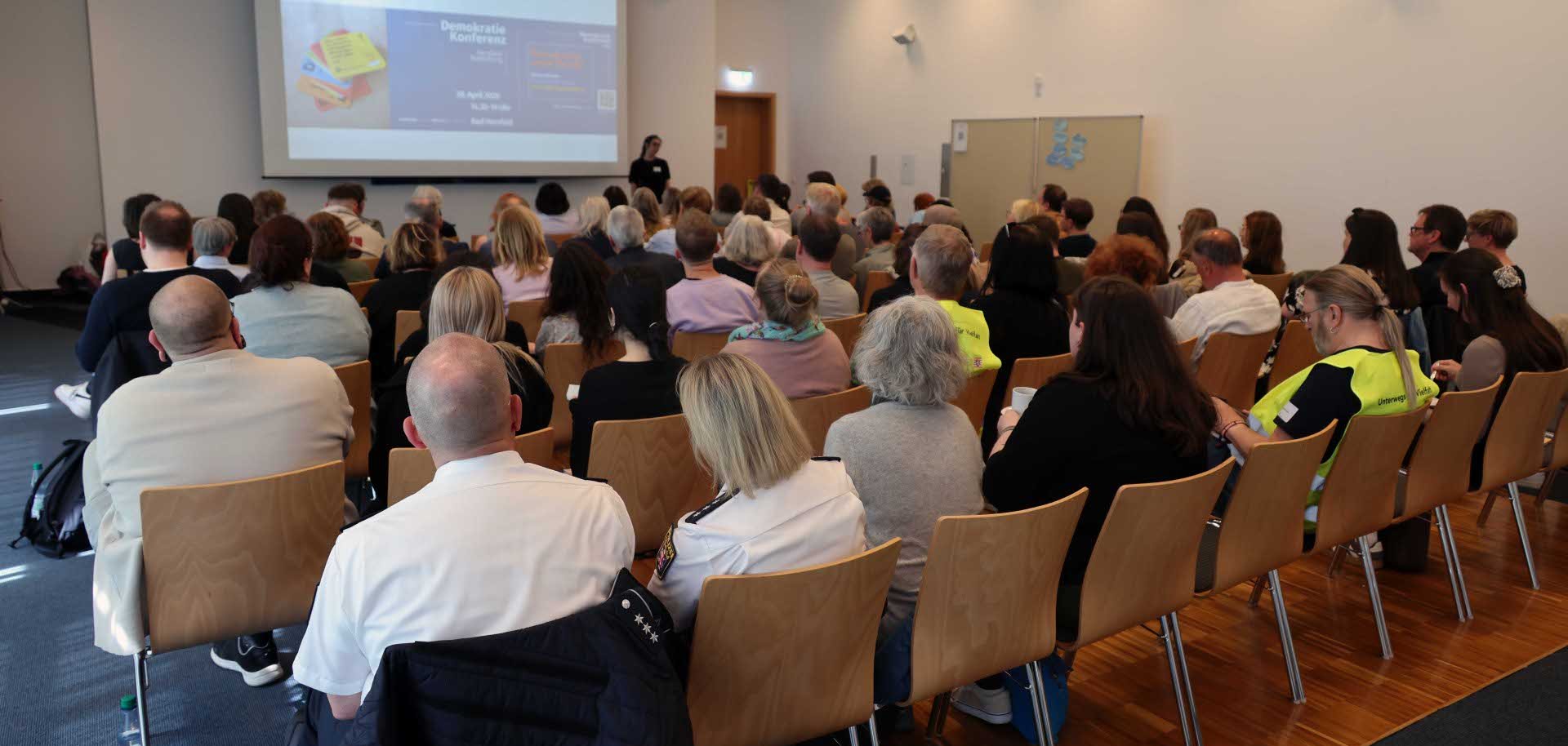 Blick in den Veranstaltungssaal der Demokratiekonferenz in der Bad Hersfelder Stadthalle. Zahlreiche Teilnehmende sitzen in Reihen vor einer großen Leinwand und verfolgen einen Vortrag. Auf der Präsentationsfolie ist die Ankündigung der Demokratiekonferenz zu sehen. Die Veranstaltung bringt Akteurinnen und Akteure aus Politik, Bildung, Zivilgesellschaft und Präventionsarbeit zusammen, um über Demokratie, gesellschaftlichen Zusammenhalt und Extremismusprävention im Landkreis Hersfeld-Rotenburg zu diskutieren.