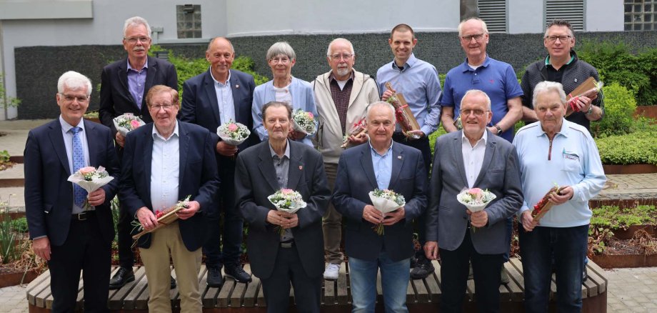 Eine Gruppe von Männern und eine Frau stehen im Innenhof einer Kreisverwaltung. Sie haben zum Teil Blumensträuße in der Hand und zum Teil eine Stracke mit einem Brettchen. Sie freuen sich.