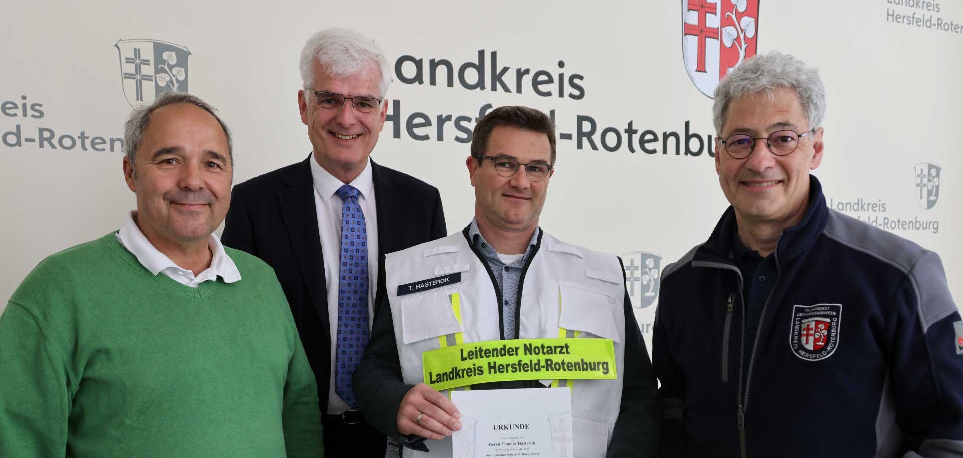 Vier Männer stehen nebeneinander vor einer Wand mit der Aufschrift „Landkreis Hersfeld-Rotenburg“. In der Mitte hält ein Mann in einer weißen Einsatzweste mit reflektierenden Streifen ein Zertifikat in den Händen; auf seiner Weste steht „Leitender Notarzt Landkreis Hersfeld-Rotenburg“. Die drei anderen Männer tragen Alltags- bzw. Dienstkleidung, einer rechts hält einen leuchtend gelben Schutzhelm. Alle blicken in die Kamera und lächeln.