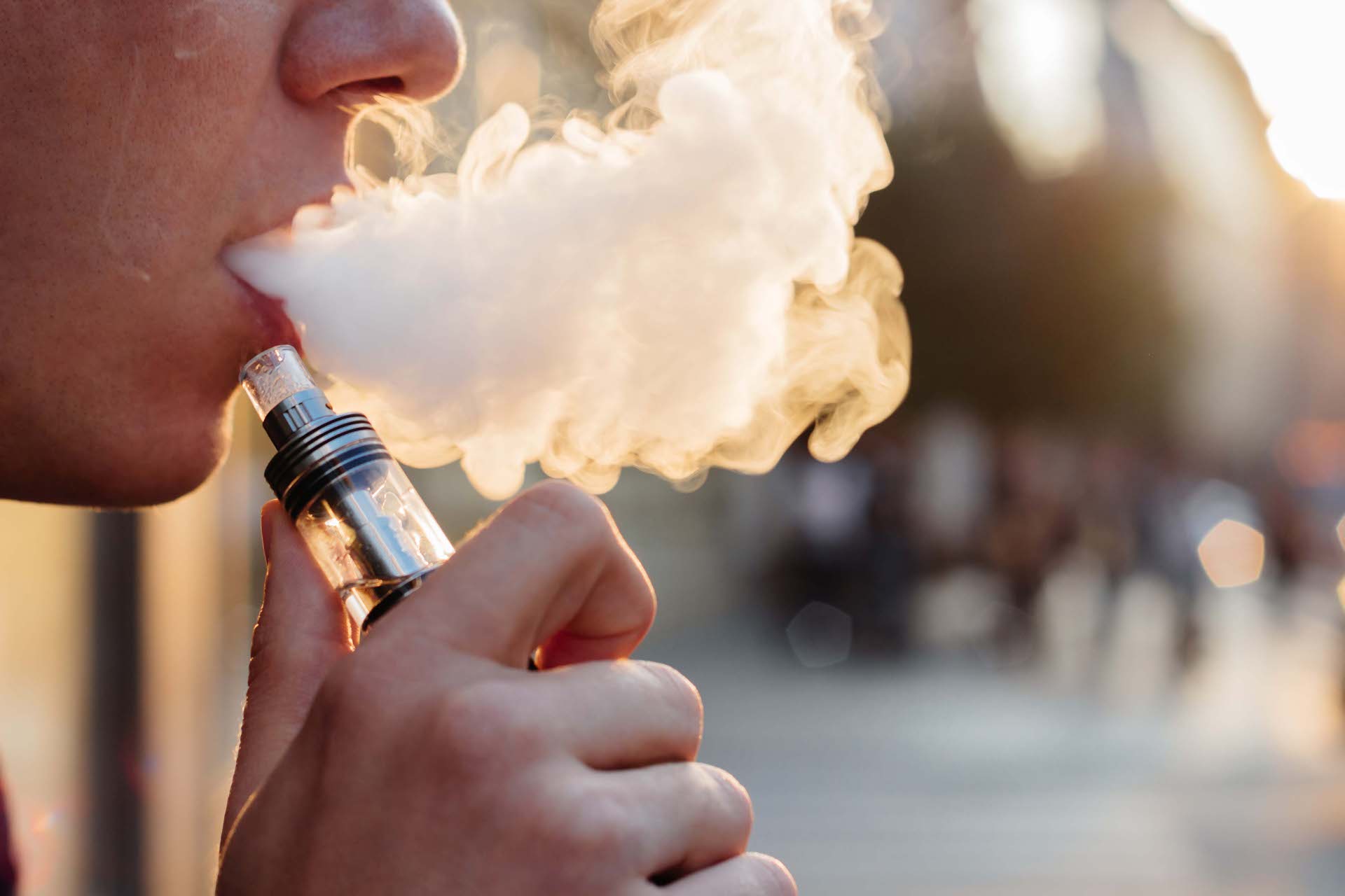 Nahaufnahme einer Person, die eine E-Zigarette (Vape) benutzt und eine dichte Wolke aus Dampf ausatmet. Die Szene ist im warmen Abendlicht aufgenommen, der Hintergrund ist unscharf.