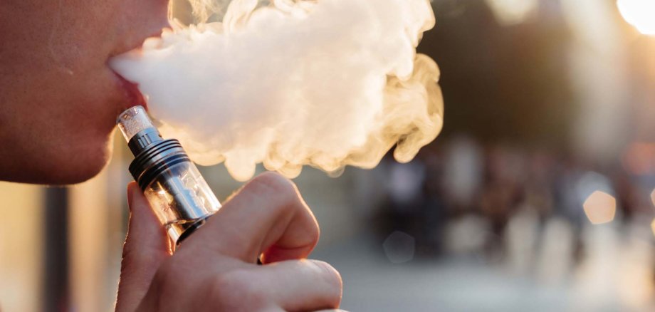 Nahaufnahme einer Person, die eine E-Zigarette (Vape) benutzt und eine dichte Wolke aus Dampf ausatmet. Die Szene ist im warmen Abendlicht aufgenommen, der Hintergrund ist unscharf.