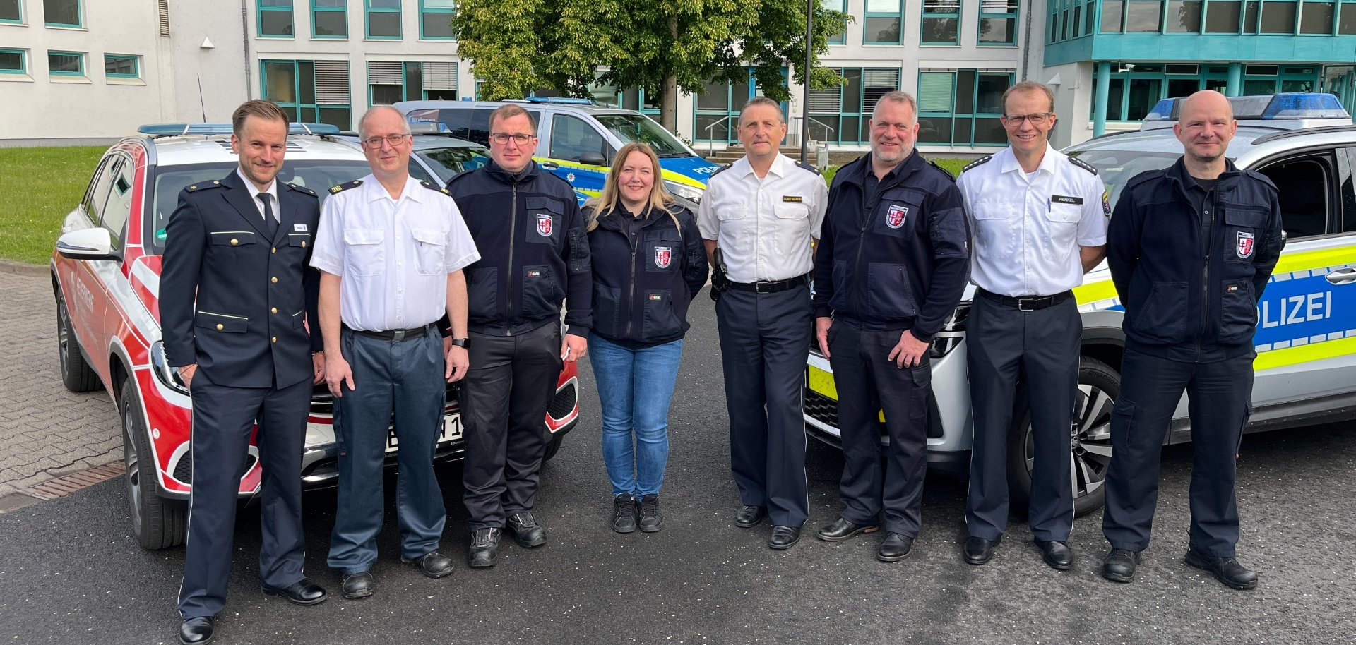 Acht Personen stehen vor zwei Polizeiautos und einem Feuerwehrwagen. Im Hintergrund sieht man ein Gebäude, die Polizeistation Bad Hersfeld.