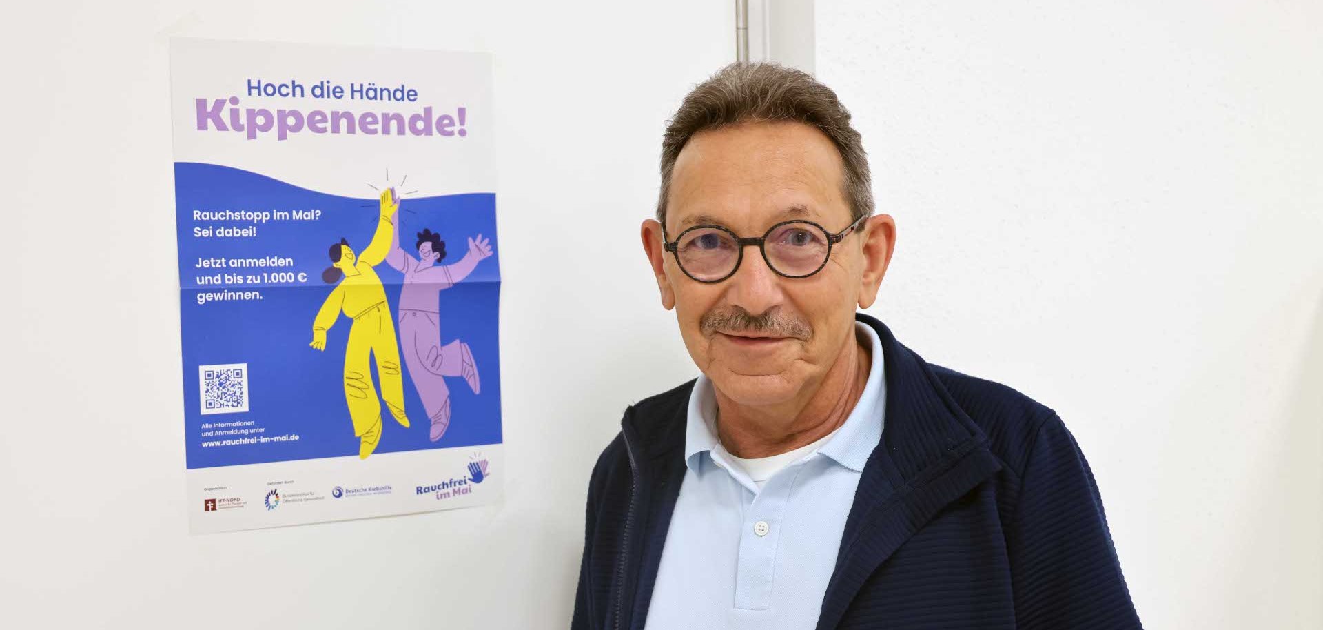 „Ein älterer Mann mit Brille und Schnurrbart steht in einem hellen Raum neben einem Poster zur Rauchstopp-Kampagne „Rauchfrei im Mai“. Er trägt ein hellblaues Poloshirt und eine dunkle Jacke und blickt in die Kamera.“