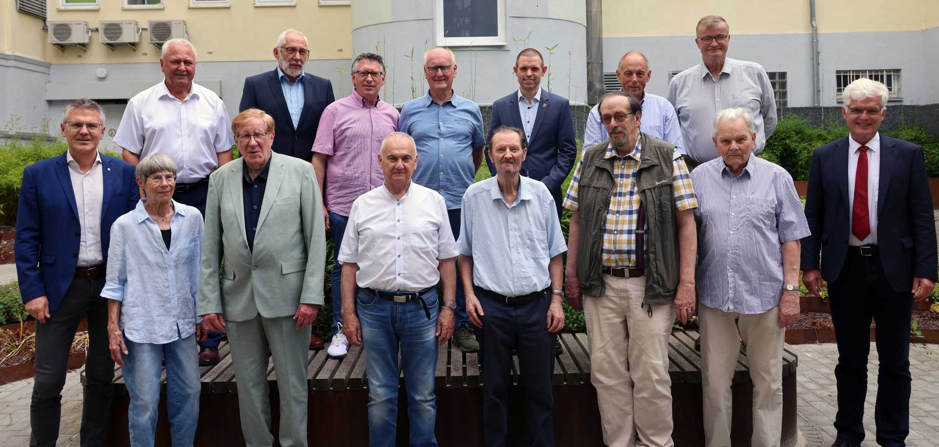 Gruppenfoto von 16 Personen unterschiedlichen Alters in einem begrünten Innenhof vor einem hellen Gebäude. Die Personen stehen in zwei Reihen und blicken in die Kamera. Einige tragen Anzug oder Sakko, andere legere Kleidung wie Hemden und Jeans. Im Hintergrund sind Fenster, Klimaanlagen und bepflanzte Beete zu sehen.