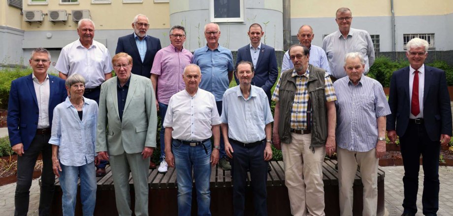 Gruppenfoto von 16 Personen unterschiedlichen Alters in einem begrünten Innenhof vor einem hellen Gebäude. Die Personen stehen in zwei Reihen und blicken in die Kamera. Einige tragen Anzug oder Sakko, andere legere Kleidung wie Hemden und Jeans. Im Hintergrund sind Fenster, Klimaanlagen und bepflanzte Beete zu sehen.