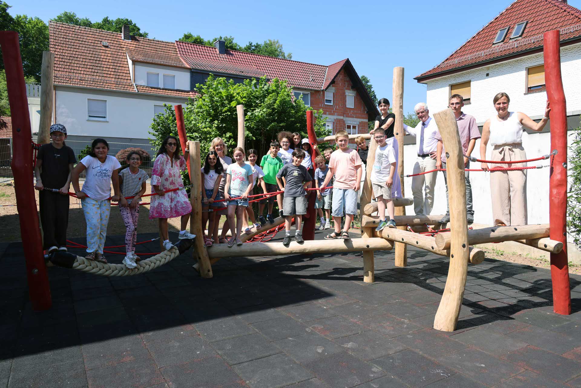 Kinder sowie Erwachsene stehen gemeinsam auf einem neuen Kletter- und Balancierparcours aus Holz und Seilen auf einem Schulhof und präsentieren die fertiggestellte Spielanlage bei sonnigem Wetter.