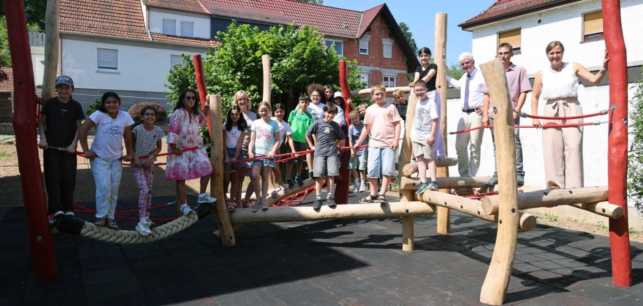 Kinder sowie Erwachsene stehen gemeinsam auf einem neuen Kletter- und Balancierparcours aus Holz und Seilen auf einem Schulhof und präsentieren die fertiggestellte Spielanlage bei sonnigem Wetter.
