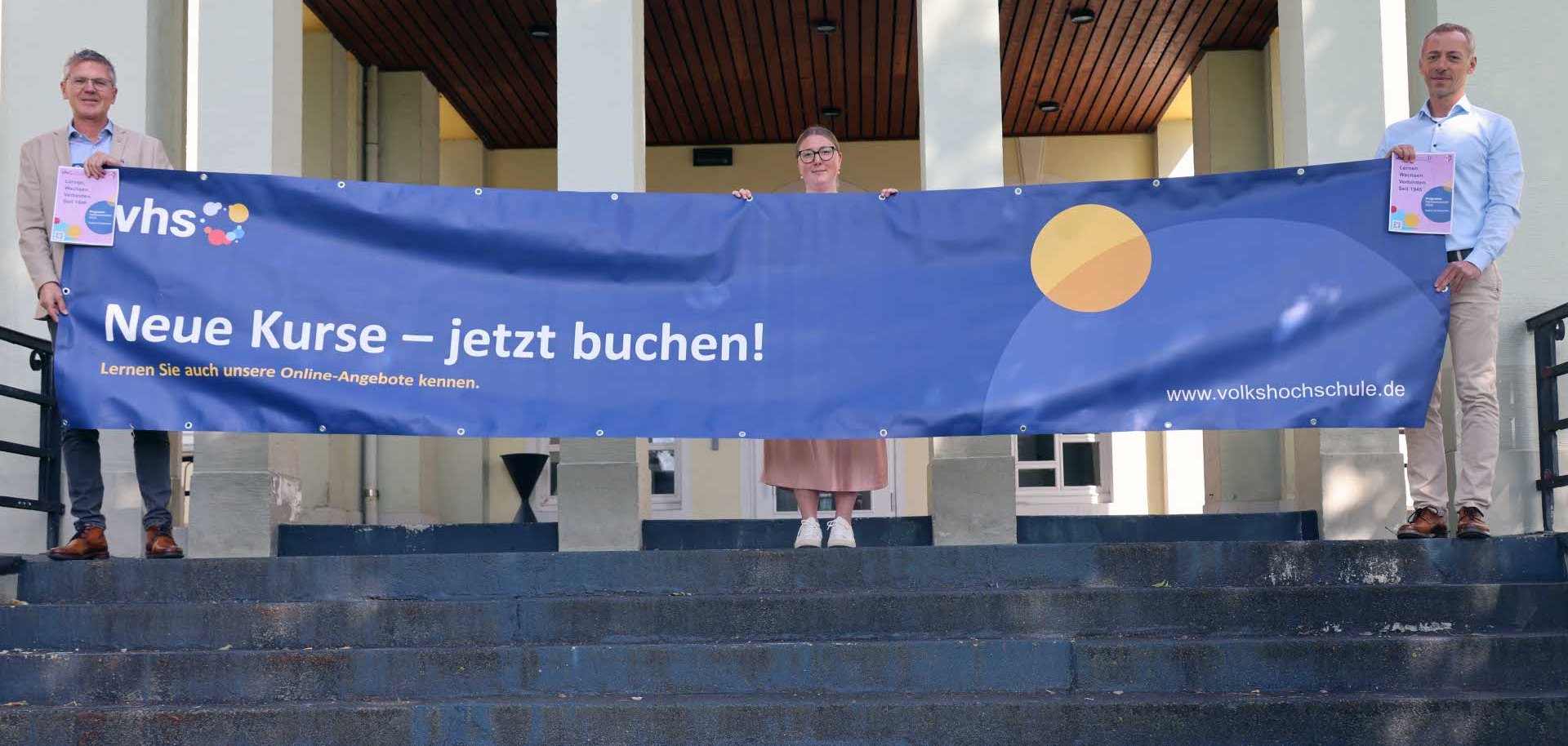 Drei Personen stehen auf einer Treppe vor einem Gebäude und halten ein großes blaues Banner der Volkshochschule (vhs) mit der Aufschrift „Neue Kurse – jetzt buchen!“. Auf dem Banner sind außerdem der Hinweis auf Online-Angebote und die Internetadresse der Volkshochschule zu sehen.