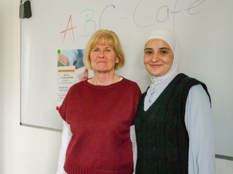 Zwei Frauen stehen lächelnd nebeneinander vor einer Tafel im ABC-Café. Das Foto zeigt eine ältere Frau im roten Oberteil und eine jüngere Frau mit weißem Kopftuch und dunkler Strickweste. Beide sind am Projekt zum Lesen- und Schreibenlernen beteiligt.