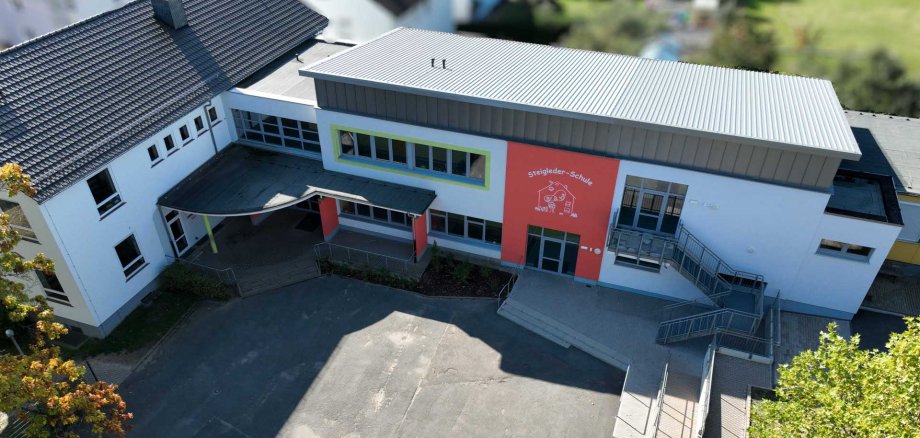 Luftaufnahme der Steigleder-Schule mit hellem Außenputz, roten und grünen Fassadenelementen, dunklem Ziegeldach und modernem Flachdach. Vor dem Gebäude befinden sich der gepflasterte Eingangsbereich mit Treppen und barrierefreien Rampen sowie ein asphaltierter Schulhof. Umgeben ist das Schulgebäude von Bäumen und Grünflächen.