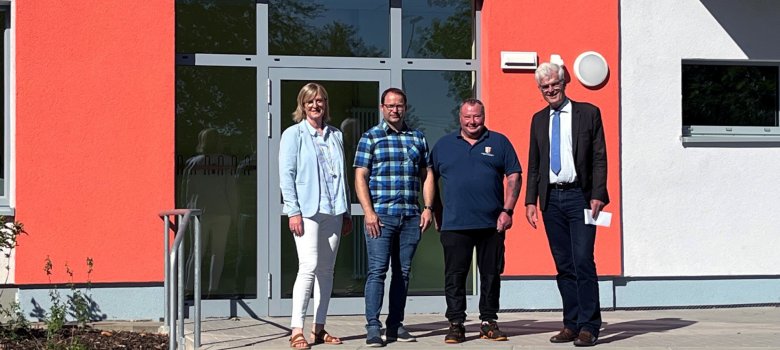 Vier Personen stehen gemeinsam vor dem Eingang der Steigleder-Schule. Sie sind unterschiedlich gekleidet, unter anderem mit heller Jacke, kariertem Hemd, Poloshirt und dunklem Anzug. Im Hintergrund sind die orangefarbene Fassade, die Glastür und die Eingangstreppe des Schulgebäudes zu sehen.
