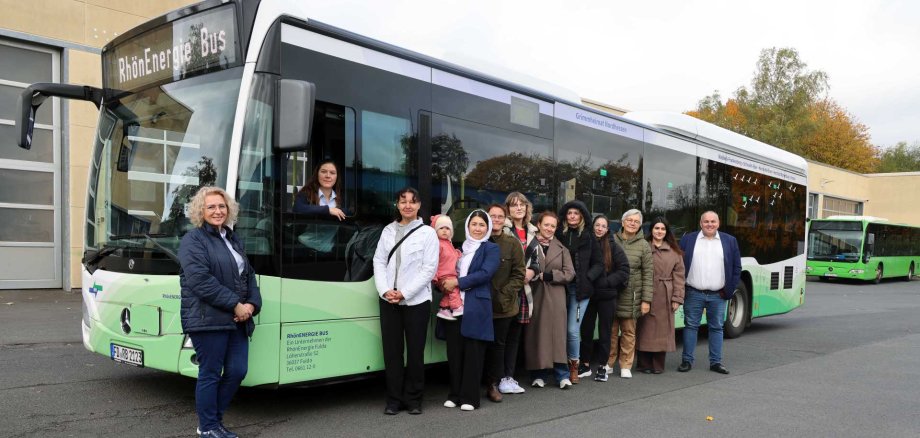 Eine Gruppe von Frauen und einem Mann steht gemeinsam mit einem Kleinkind vor einem grün-weißen Linienbus der RhönEnergie Bus. Eine weitere Frau schaut aus dem geöffneten Fahrerfenster. Die Aufnahme entstand auf einem Betriebsgelände vor einem Gebäude. Im Hintergrund ist ein weiterer grüner Bus zu sehen.