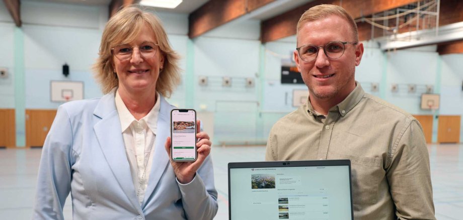 Zwei Personen präsentieren in einer Sporthalle eine digitale Plattform auf einem Smartphone und einem Laptop. Die Frau trägt einen hellblauen Blazer, der Mann ein olivgrünes Hemd. Im Hintergrund sind Basketballkörbe und Sportgeräte zu sehen.
