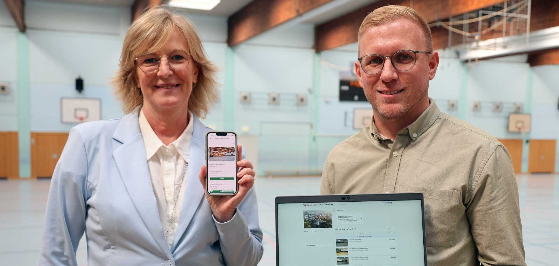 Zwei Personen präsentieren in einer Sporthalle eine digitale Plattform auf einem Smartphone und einem Laptop. Die Frau trägt einen hellblauen Blazer, der Mann ein olivgrünes Hemd. Im Hintergrund sind Basketballkörbe und Sportgeräte zu sehen.