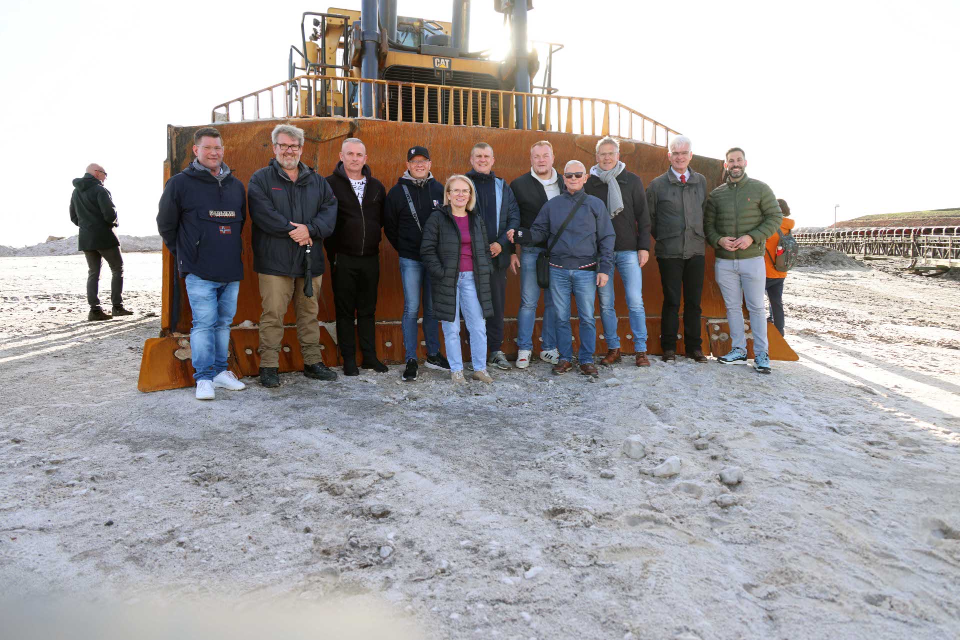 11 Personen stehen vor einem großen Bagger und schauen in die Kamera. Im Vordergrund ist ein weißer Untergrund zu erkennen.