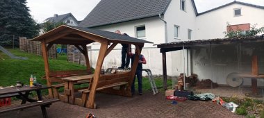Mehrere Personen arbeiten an einem großen hölzernen Unterstand mit Sitzbänken. Eine Person steht auf einer Leiter am Dach, während Werkzeuge und Arbeitsmaterialien auf dem Boden liegen.