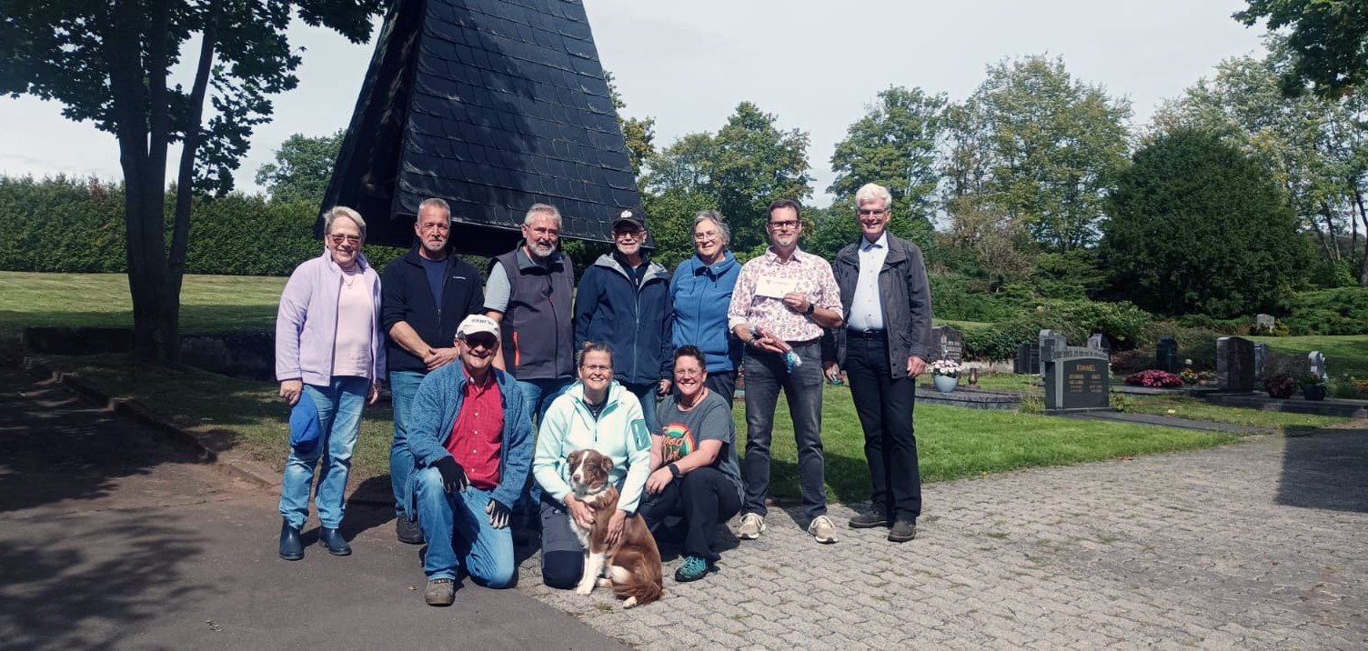 Gruppe von Erwachsenen steht bei einer gemeinsamen Aufräum- oder Arbeitseinsatzaktion im Grünen. Einige Personen tragen Arbeitskleidung und halten Gartengeräte.