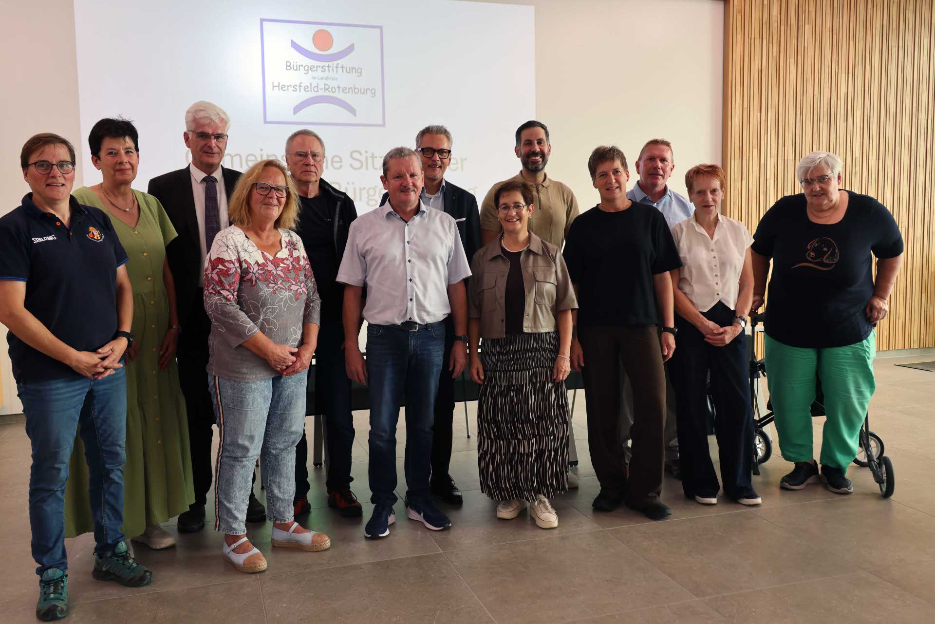 Gruppenfoto mit 14 Personen, die gemeinsam in einem Veranstaltungsraum vor einer Leinwand stehen. Auf der Leinwand ist das Logo der Bürgerstiftung Hersfeld-Rotenburg zu sehen. Die Personen stehen in zwei Reihen und blicken in die Kamera. Im Hintergrund sind außerdem helle Wände und eine Holzverkleidung zu sehen.