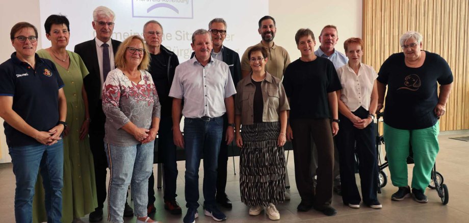 Gruppenfoto mit 14 Personen, die gemeinsam in einem Veranstaltungsraum vor einer Leinwand stehen. Auf der Leinwand ist das Logo der Bürgerstiftung Hersfeld-Rotenburg zu sehen. Die Personen stehen in zwei Reihen und blicken in die Kamera. Im Hintergrund sind außerdem helle Wände und eine Holzverkleidung zu sehen.