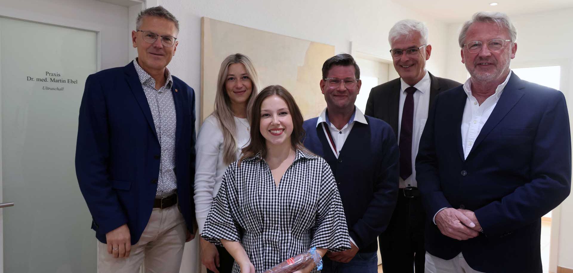 Auf dem Foto sind insgesamt sechs Personen zu sehen. Auf der linken Seite ist eine Tür mit einer Aufschrift zu erkennen. Im Hintergrund befindet sich ein großes Bild.