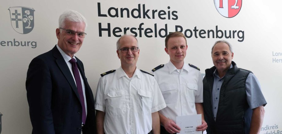 Vier Männer stehen für ein Gruppenfoto vor einer Wand mit dem Schriftzug „Landkreis Hersfeld-Rotenburg“. Zwei Männer tragen weiße Uniformhemden, einer hält eine Urkunde in den Händen. Die beiden anderen Männer tragen einen dunklen Anzug beziehungsweise eine dunkle Weste über einem Hemd. Alle blicken in die Kamera und lächeln.