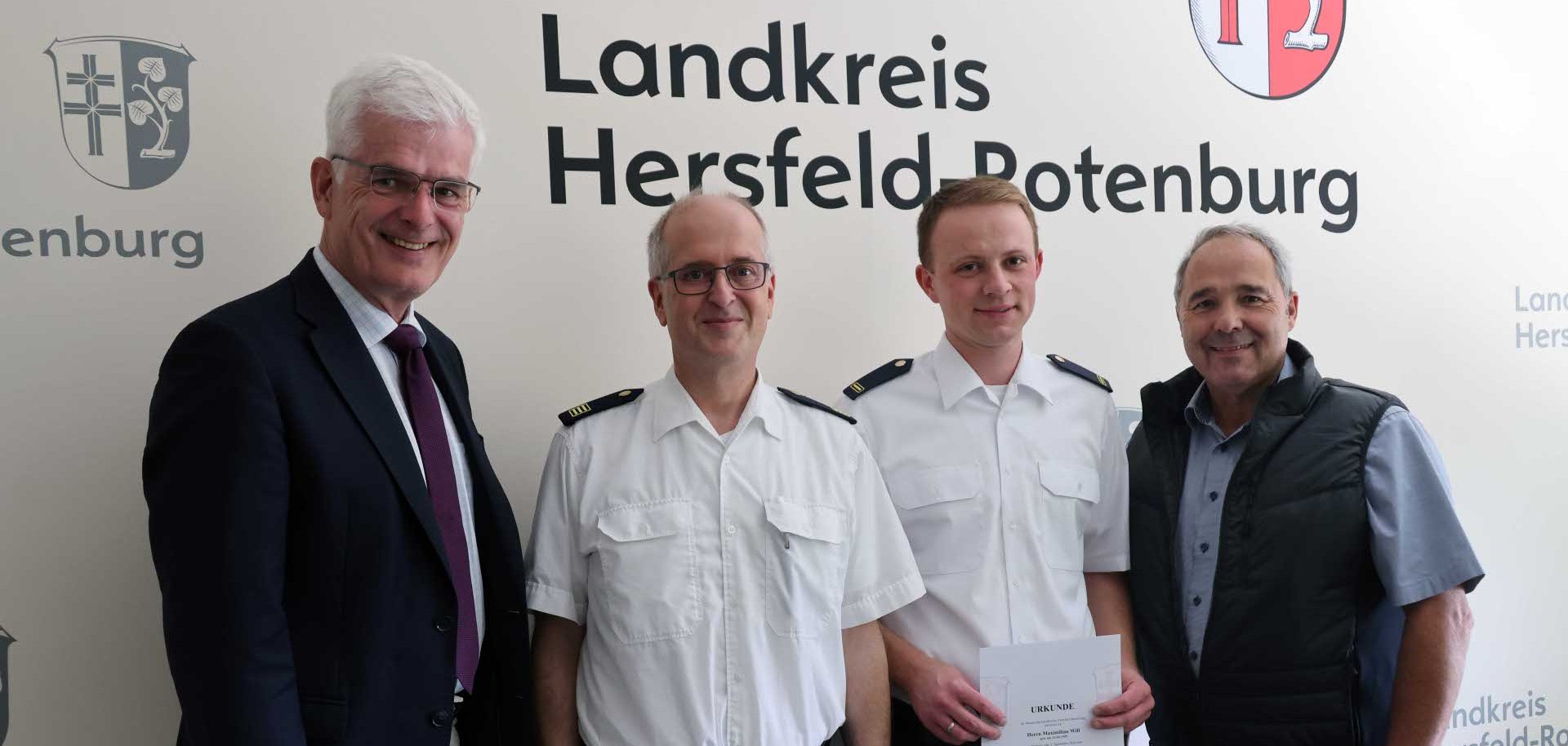 Vier Männer stehen für ein Gruppenfoto vor einer Wand mit dem Schriftzug „Landkreis Hersfeld-Rotenburg“. Zwei Männer tragen weiße Uniformhemden, einer hält eine Urkunde in den Händen. Die beiden anderen Männer tragen einen dunklen Anzug beziehungsweise eine dunkle Weste über einem Hemd. Alle blicken in die Kamera und lächeln.