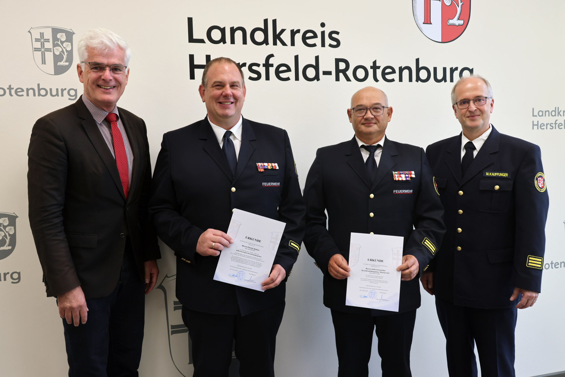 Erfahrung übergeben, Verantwortung übernommen: Landrat Torsten Landrat Torsten Warnecke, Patrick Kniest, Martin Orf und Kreisbrandinspektor Marco Kauffunger stehen vor dem Schriftzug des Landkreises. Kniest und Orf halten ihre Urkunden in den Händen.