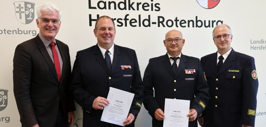 Erfahrung übergeben, Verantwortung übernommen: Landrat Torsten Landrat Torsten Warnecke, Patrick Kniest, Martin Orf und Kreisbrandinspektor Marco Kauffunger stehen vor dem Schriftzug des Landkreises. Kniest und Orf halten ihre Urkunden in den Händen.