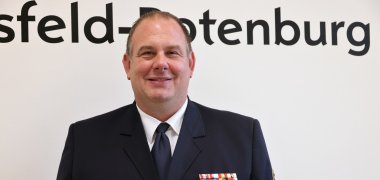 Stellvertretender Kreisbrandinspektor Patrick Kniest Patrick Kniest lächelt in dunkelblauer Feuerwehruniform vor dem Schriftzug des Landkreises Hersfeld-Rotenburg in die Kamera.