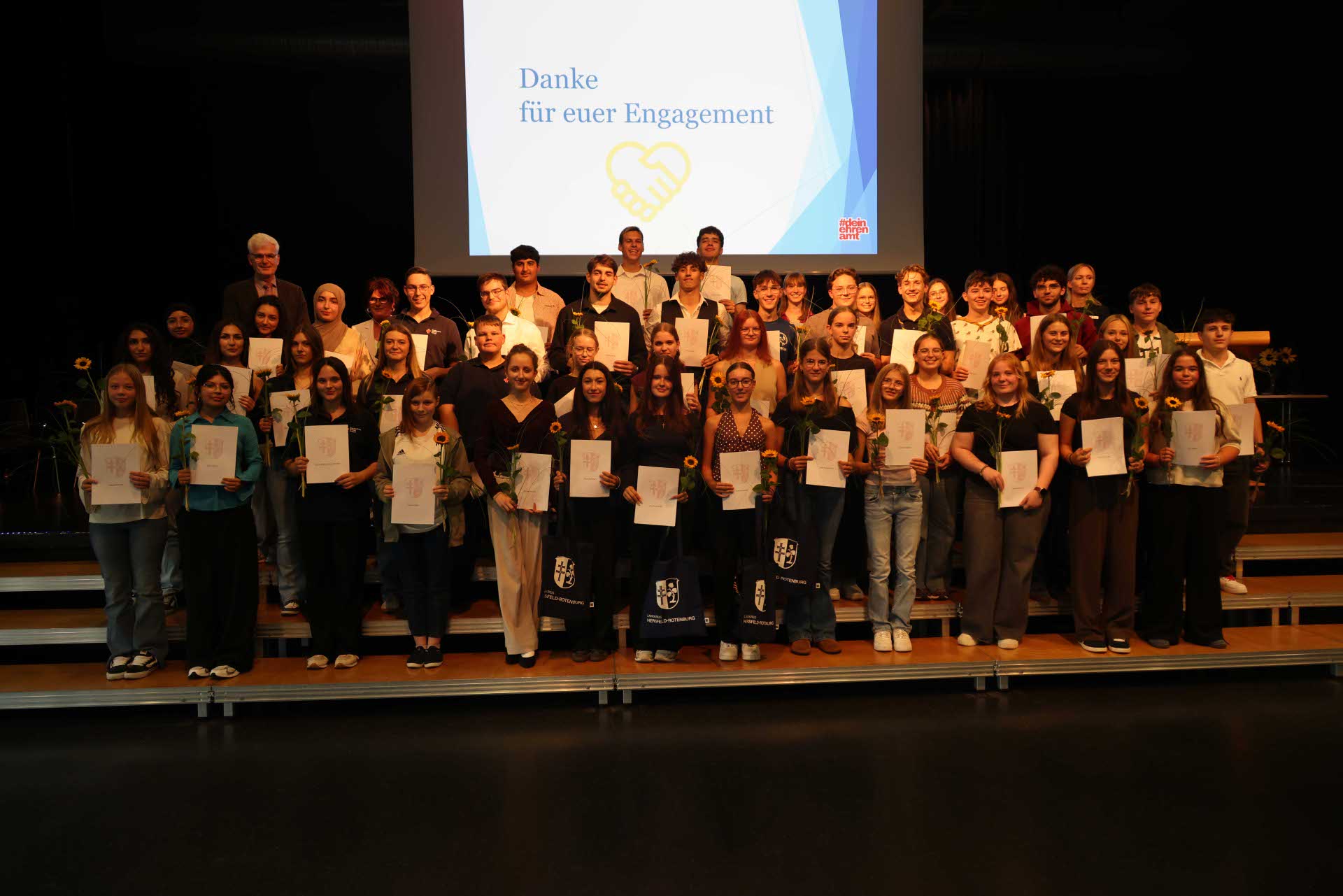 Gruppenfoto mit zahlreichen Jugendlichen und Erwachsenen, die gemeinsam auf einer Bühne stehen und Urkunden sowie Sonnenblumen halten. Im Hintergrund ist eine Leinwand mit der Aufschrift ‚Danke für euer Engagement‘ zu sehen.“