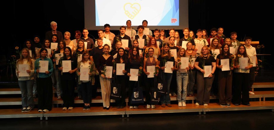 Gruppenfoto mit zahlreichen Jugendlichen und Erwachsenen, die gemeinsam auf einer Bühne stehen und Urkunden sowie Sonnenblumen halten. Im Hintergrund ist eine Leinwand mit der Aufschrift ‚Danke für euer Engagement‘ zu sehen.“